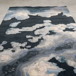A rectangular area rug with an abstract cloud-like pattern in shades of navy, charcoal, light blue, and cream, laid out on a light gray floor.