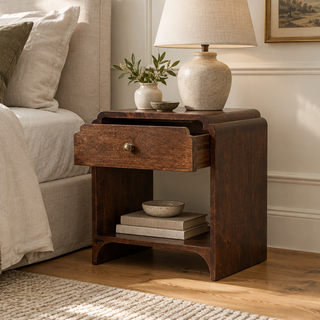 Dark wood bedside table with a stepped curved top, open drawer and lower shelf holding books, beside a linen-covered bed.