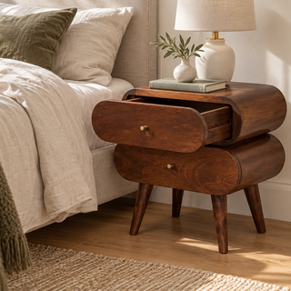 Rounded dark wood nightstand with two drawers and brass knobs, top drawer open, beside a cream bed with a ceramic lamp.