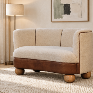 A modern two-seater sofa with cream bouclé upholstery, a brown leather base, and round wooden legs, styled in a warm, neutral-toned living room setting.