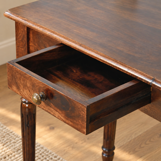 Close-up of an open wooden table drawer with a round brass knob, showing dark wood grain, a rounded tabletop edge and turned legs.