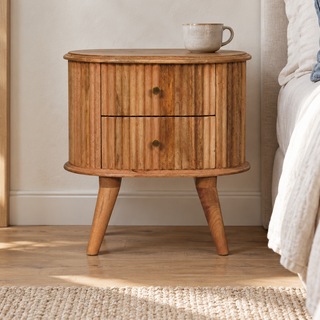 A round wooden nightstand with two drawers and angled legs, styled in a bedroom next to a bed with white bedding and a mug on top.