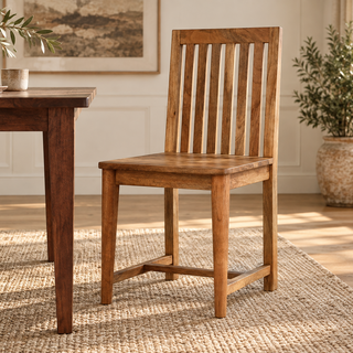 A wooden dining chair with a slatted back and straight legs, shown in a bright dining room next to a matching wooden table on a woven rug.
