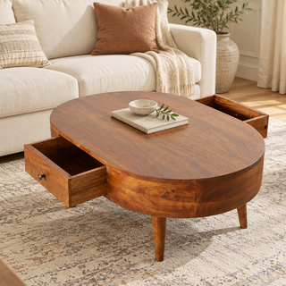 An oval mid-century modern wooden coffee table with two pull-out storage drawers, shown in a bright, modern living room next to a cream sofa.