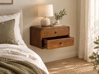 A floating wooden nightstand with two drawers and small round brass knobs, styled in a bedroom next to a bed with white linens. A textured ceramic table lamp and a small potted plant sit on top, with warm sunlight streaming in from a window with sheer curtains.