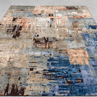 Rectangular abstract area rug with a distressed, painterly pattern in beige, light grey, terracotta and dark brown, with deep blue and periwinkle patches toward one corner, laid flat on a light grey floor.