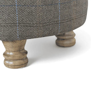 Close-up of an upholstered ottoman or stool with a brown and gray herringbone patterned fabric and turned wooden legs on a white background.